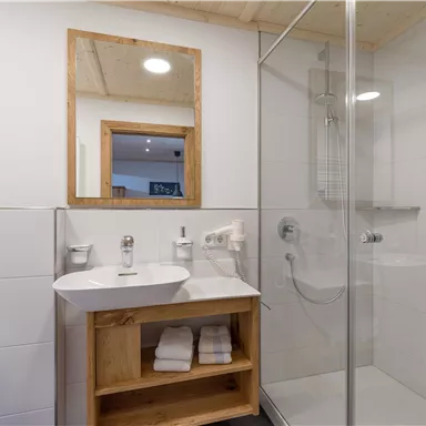 Ein modernes Badezimmer mit einem Waschbecken und einem Spiegel. Die Dusche ist mit Glaswänden getrennt und Handtücher sind ordentlich gestapelt.