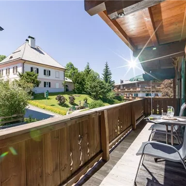 Ein Balkon mit Holzgeländer und zwei Stühlen. Im Hintergrund sieht man ein grünes Grundstück und Häuser unter klarem Himmel.