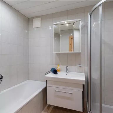 Ein modernes Badezimmer mit einer Badewanne und einer Dusche. Die Wände sind hell gefliest, und es gibt einen Spiegel über dem Waschbecken.
