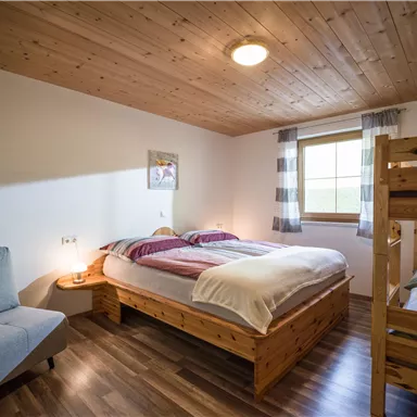 Ein gemütliches Schlafzimmer mit einem Doppelbett und einem Etagenbett. Der Raum hat Holzverkleidung und ein Fenster, das Tageslicht hereinlässt.