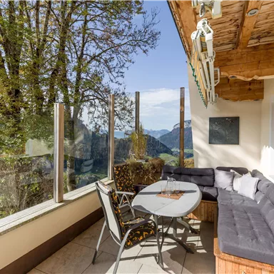 Eine gemütliche Terrasse mit Sitzmöglichkeiten und einem Tisch. Im Hintergrund sind Bäume und Berge zu sehen, unter einem klaren Himmel.