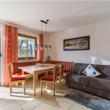 Ein gemütlicher Essbereich mit einem Tisch und Stühlen, umgeben von Fenstern und hellen Vorhängen. Auf der rechten Seite befindet sich ein bequemer grauer Sofa mit dekorativen Kissen.