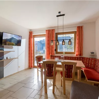 Ein gemütlicher Essbereich mit einem großen Tisch und roten Sitzpolstern. Große Fenster lassen viel Licht herein und bieten einen schönen Blick auf die Berge.
