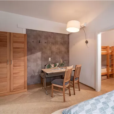 A cozy room with a wooden table and chairs. On the left, there is a wardrobe, and on the right, there is an access to another room with a bunk bed.