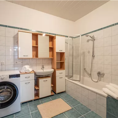 A modern bathroom with a bathtub and a shower. Next to the bathtub is a washing machine, and fresh towels are visible.