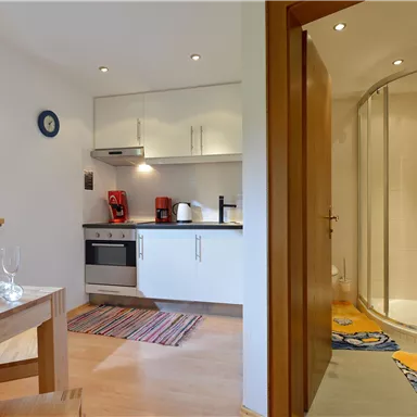 A modern kitchen with white cabinets and a dining area. In the background, a bathroom with a shower stall is visible.