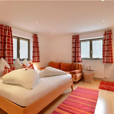 A bright bedroom with a large bed and a cozy sofa. Red curtains and a carpet along with natural light create an inviting atmosphere.