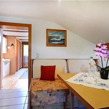 A cozy room with wooden furniture and a decoration featuring an orchid. In the background, an open space and a picture with a landscape inspiration are visible.