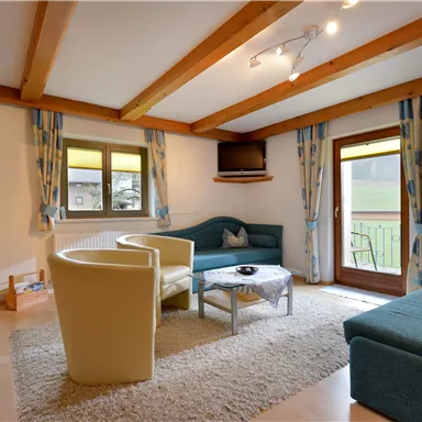 A cozy living room with wooden beams and modern furniture. There is a sofa, armchair, and a table in front of a window with a view outside.