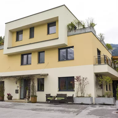 Ein modernes zweigeschossiges Haus in einer grünen Umgebung. Die Fassade ist gelb und hat große Fenster.