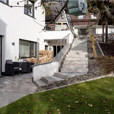 Ein modernes Haus mit einer Terrasse und einem gepflasterten Weg. Eine Treppe führt zum oberen Bereich, umgeben von Grünflächen und Bäumen.
