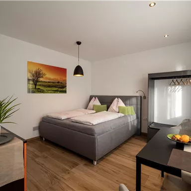 Ein modernes Schlafzimmer mit einem Doppelbett, einer kleinen Sitzecke und einem Tisch. Die Wände sind hell gestrichen und ein Kunstwerk ist an der Wand angebracht.
