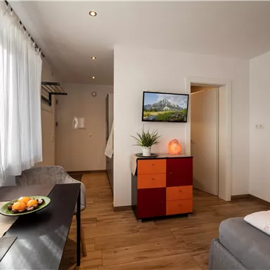 Ein modernes Zimmer mit hellen Vorhängen und einem großen Fenster. Es gibt einen Essbereich, einen Fernseher an der Wand und eine gemütliche Sitzecke.