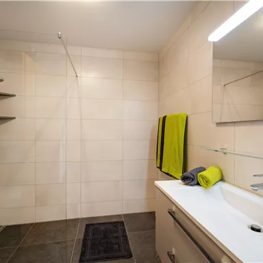 Ein modernes Badezimmer mit einer begehbaren Dusche und hellen Fliesen. Auf dem Waschbecken liegen farbige Handtücher neben einem Spiegel.
