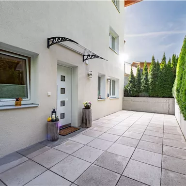 Ein heller Eingangsbereich mit einer großen Terrasse aus Fliesen. Umgeben von grünen Pflanzen und einem modernen Eingangsbereich.