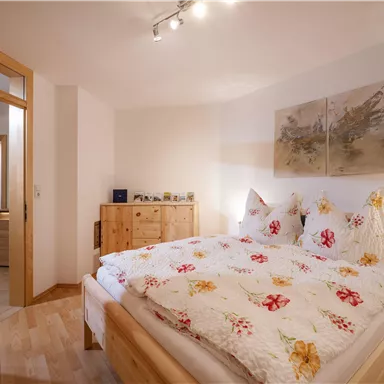 Ein gemütliches Schlafzimmer mit einem großen Bett und blumiger Bettwäsche. Im Hintergrund sind Holzkommoden und ein hübsches Wandbild zu sehen.