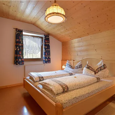A cozy bedroom with two beds and wooden ceilings. Large windows with colorful curtains let in plenty of light.