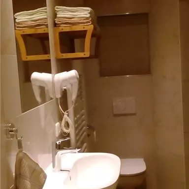 A modern bathroom with a wooden ceiling and warm beige tones. It features a sink, a toilet, and folded towels on a shelf.
