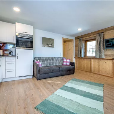Ein gemütlicher Wohnraum mit moderner Küche und einem Sofa. Der Raum ist hell und freundlich eingerichtet, mit einem Teppich und Holzelementen.