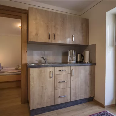 Eine gemütliche Küchenzeile mit Holzschränken und einem kleinen Waschbecken. Im Hintergrund ist ein Schlafzimmer mit Betten und einem Fenster zu sehen.