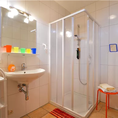 Ein modernes Badezimmer mit einer Duschkabine, einem Waschbecken und einem großen Spiegel. Die Wände sind weiß gefliest, und bunte Behälter bringen einen Farbtupfer in den Raum.