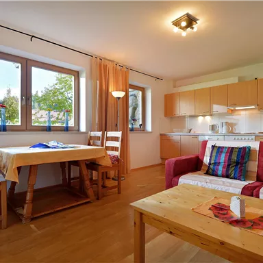 Ein helles Wohnzimmer mit einer offenen Küche. Es gibt einen Tisch mit Stühlen und eine gemütliche Sitzecke mit bunten Kissen.