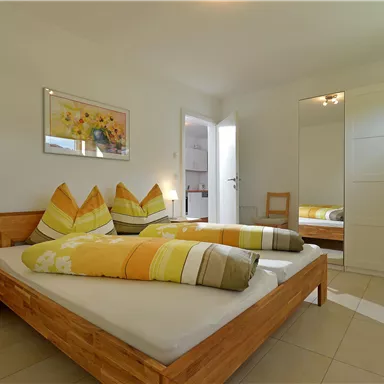 Ein helles Schlafzimmer mit zwei Betten und bunten Kissen. Im Hintergrund sind ein Schrank und eine Tür zu sehen.