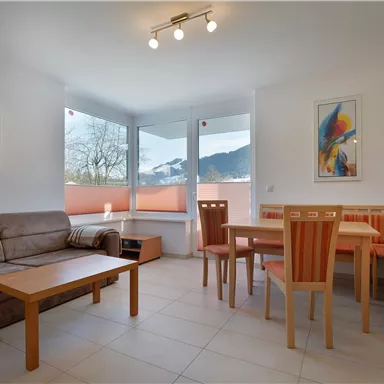 Ein helles Wohnzimmer mit einem Sofa, einem Tisch und Stühlen. Große Fenster bieten einen Blick auf die Berge.