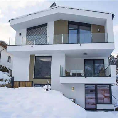 Ein modernes Haus umgeben von Schnee. Die großen Fenster bieten einen Blick auf die winterliche Landschaft.