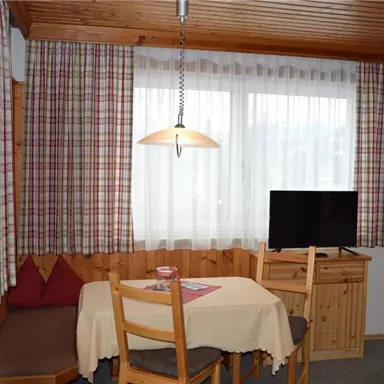 A cozy room with a dining table and chairs. Large windows let in plenty of light, and a television is placed in the corner.