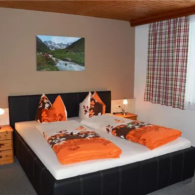 A cozy bedroom with a double bed, decorated in orange and white. A painting of a mountain landscape hangs on the wall, and a window with curtains lets natural light in.