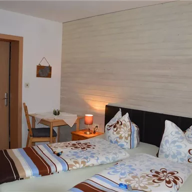 A bright bedroom with two single beds and colorful bed covers. There is a small table and a chair next to the door.