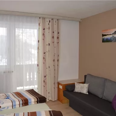 A cozy room with two beds and a gray sofa. Large windows with curtains provide plenty of daylight.