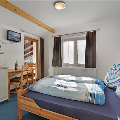 A cozy room with a double bed and colorful cushions. There is a small dining table, a kitchenette, and a window with a view outside.