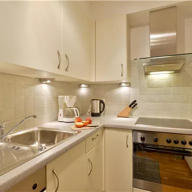 A modern kitchenette with bright cabinets and stainless steel appliances. On the countertop are a coffee machine and some fruits.