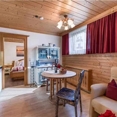 Eine gemütliche Wohnküche mit Holzwänden und einem runden Tisch. Im Hintergrund sind ein Schlafzimmer und dekorative Möbel zu sehen.