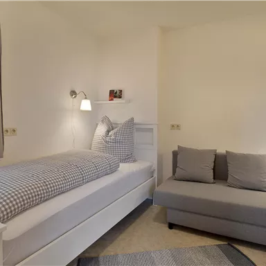 A simple bedroom with a single bed and a gray sofa. The walls are light, and there are curtains at a window.