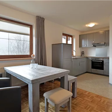 A modern kitchen corner with a table and chairs. The walls are bright and the windows provide plenty of daylight.