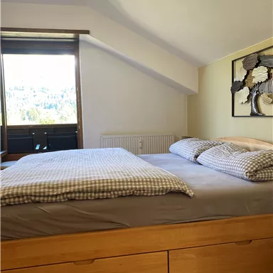 Ein gemütliches Schlafzimmer mit einem großen Bett und einem Fenster, das auf die Natur blickt. Die Wände sind hell und es gibt dekorative Elemente im Raum.