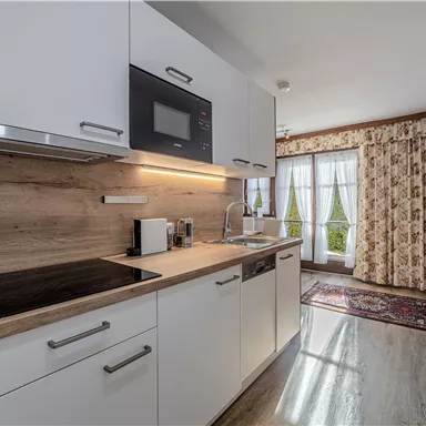Eine moderne Küche mit weißen Schränken und Holzregalen. Durch die Fenster scheint Tageslicht und sorgt für eine freundliche Atmosphäre.