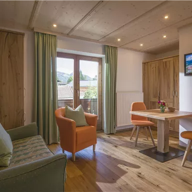 Ein modernes Wohn- und Esszimmer mit Holzboden und großen Fenstern. Die gemütliche Einrichtung umfasst ein Sofa, zwei Stühle und einen Tisch, umgeben von sanften Farbtönen.