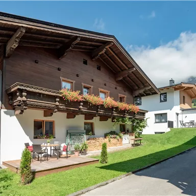A charming wooden house in the mountains with blooming balcony plants. In the background, snow-covered peaks and a clear sky are visible.