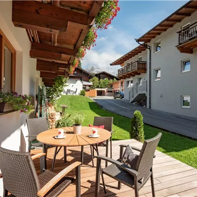 An inviting terrace with a table and chairs, surrounded by green grass. In the background, modern buildings and a sunny landscape can be seen.