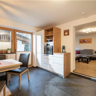 A modern kitchen with a dining table and chairs. In the background, a living room can be seen, which is cozily furnished.