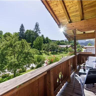 A beautiful balcony with a wooden railing and two chairs. In the background, green trees and a sunny landscape can be seen.