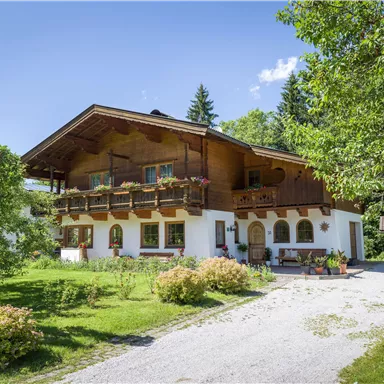 A beautiful alpine house with wooden decorations and a balcony. The garden is green and blooming, surrounded by trees.