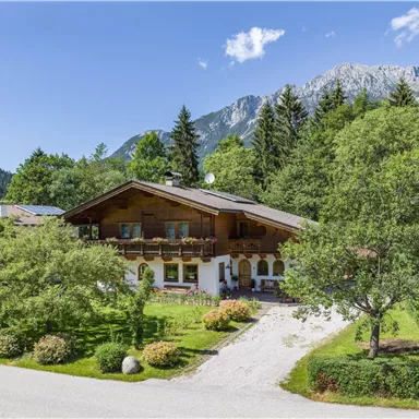 A picturesque house surrounded by trees and green spaces. Majestic mountains and a clear sky can be seen in the background.