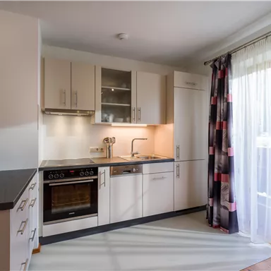 A modern kitchen with light furniture and a large window front. The room is well-lit and inviting.