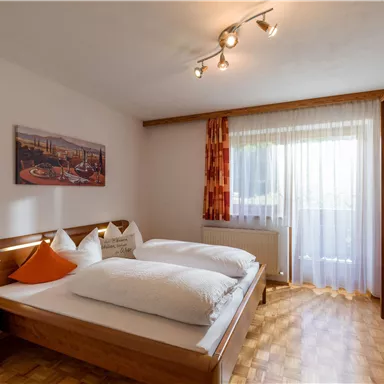 A cozy bedroom with a double bed and light wooden furniture. Large windows let in plenty of daylight.