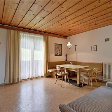 A cozy living space with a wooden ceiling and a dining table. The windows let in plenty of natural light, and the furnishings are simple and inviting.